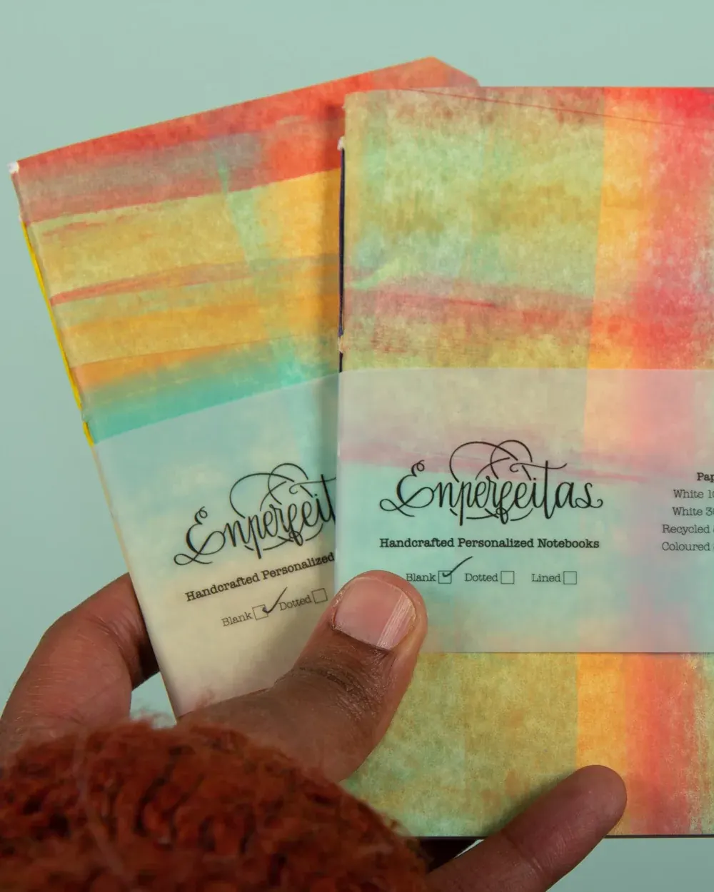 Close-up of a hand holding two watercolor-designed notebooks with the brand name 'Emperletas' on the covers. The notebooks are labeled as handcrafted personalized notebooks, with options for blank, dotted, or lined pages checked off.
